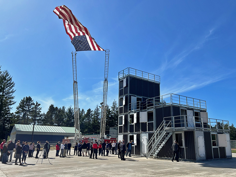 a united states flag flies between two fire truck ladders next to a four-story steel fire training tower. a crowd of people is around the building.