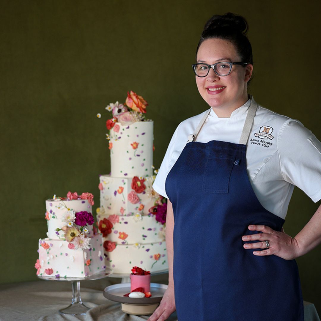 Photo of Pastry Chef Alison Murphy standing in front of table with decorated tiered cakes sitting on it